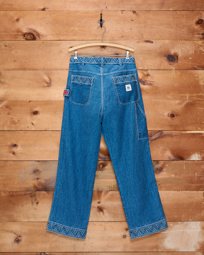 Embroidered Denim Knolly Brook Trousers displayed on a wooden wall, showcasing the back with distinct pockets and white stitching. These classic cotton trousers blend durable workwear construction with timeless style.