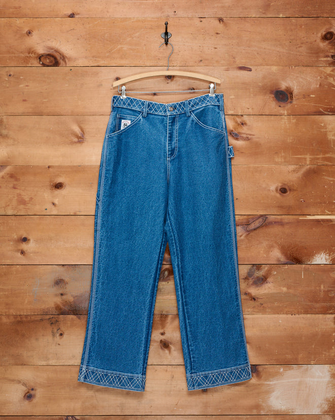The Embroidered Denim Knolly Brook Trousers, featuring white stitching reminiscent of classic workwear, hang on a wooden background from a hanger and wall hook.