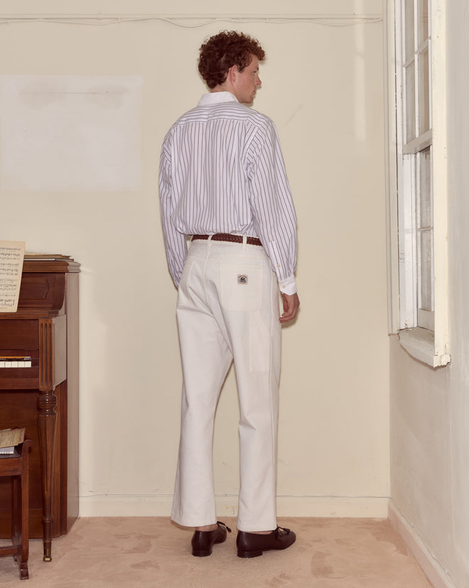 A man stands facing a corner in a room, wearing a striped shirt and crisp white Twill Knolly Brook Trousers.