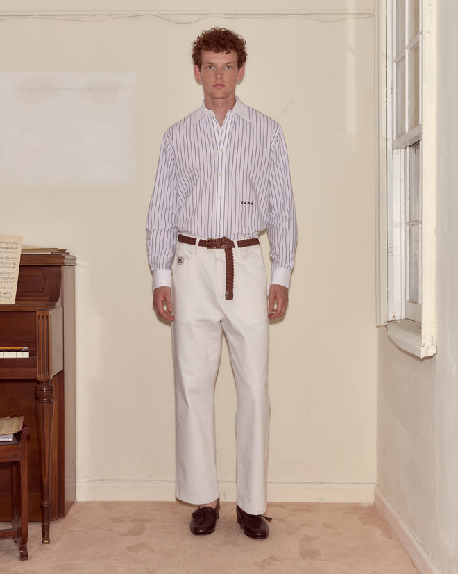 A man stands indoors by a piano wearing a striped shirt, white Twill Knolly Brook Trousers, and a brown belt.