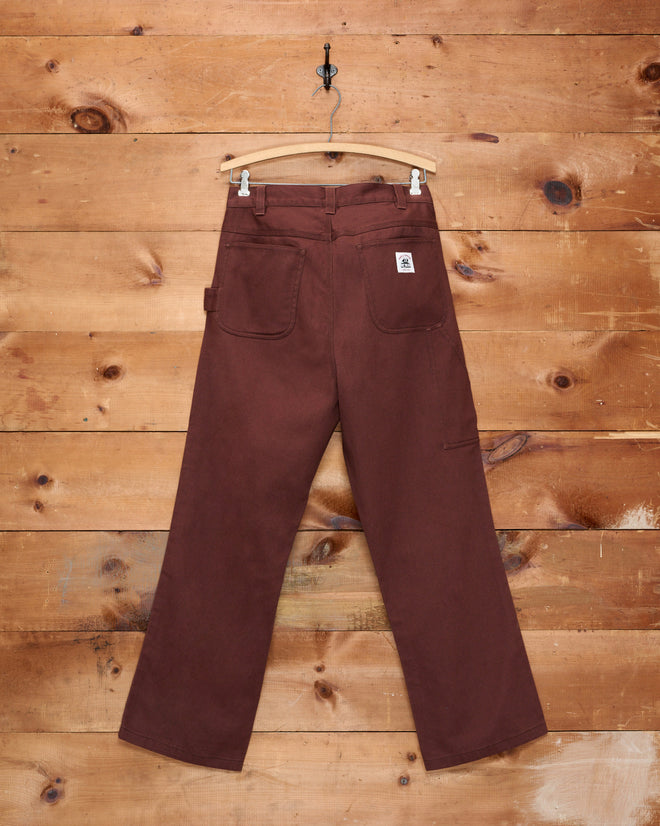 The Twill Knolly Brook Trousers - Brown, featuring wide legs and crafted from cotton twill, hang on a wooden hanger against a rustic wood wall.