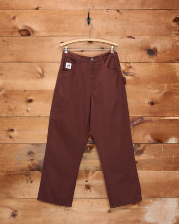 The Twill Knolly Brook Trousers in brown, displayed on a wooden hanger against a rustic wooden wall background.