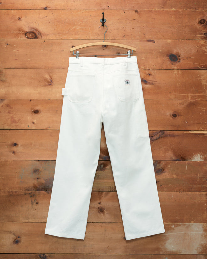 The Twill Knolly Brook Trousers in white hang on a wooden hanger against a rustic wooden wall, showcasing their classic workwear-inspired design reminiscent of Bode’s aesthetic.