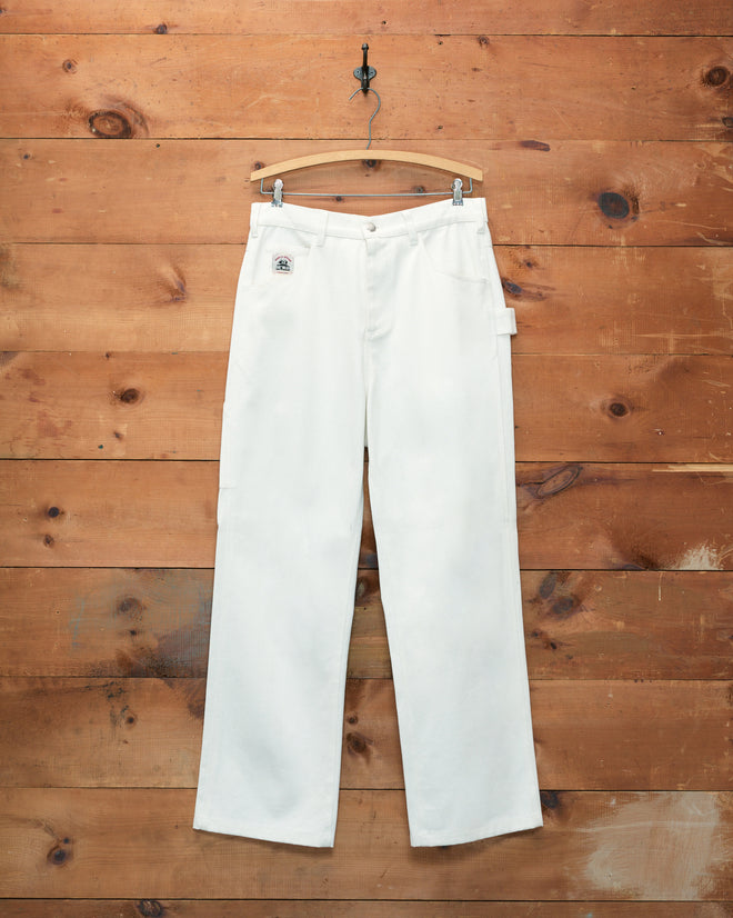 Twill Knolly Brook Trousers in white cotton twill hang on a wooden hanger against a wooden wall, evoking classic workwear style.