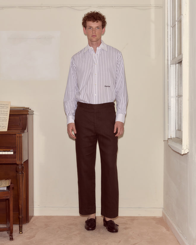 A man stands indoors beside a piano wearing a striped shirt, loafers, and Lightweight Linen Side-Buckle Trousers in Brown/Black.