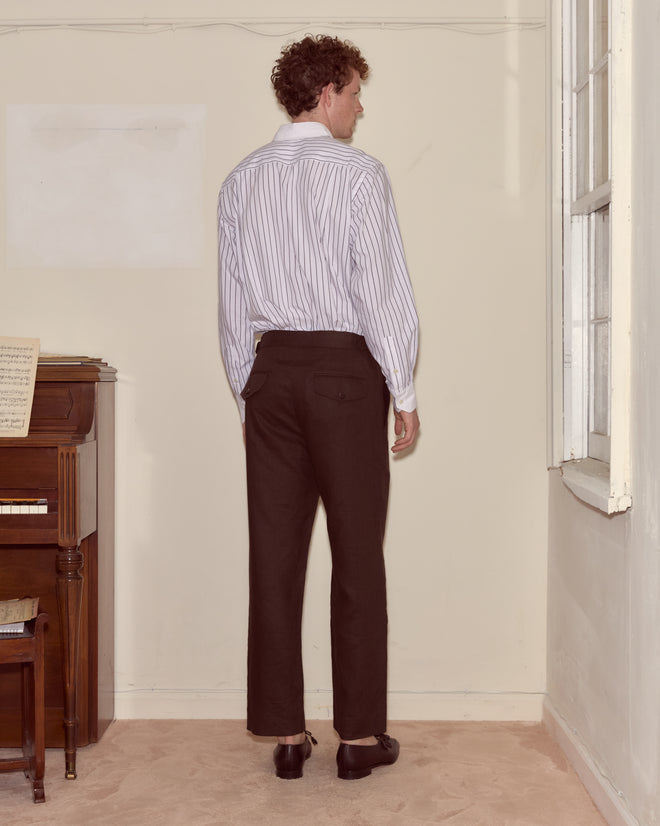 A man in a striped shirt and Lightweight Linen Side-Buckle Trousers - Brown/Black stands facing a wall beside a piano and a window.