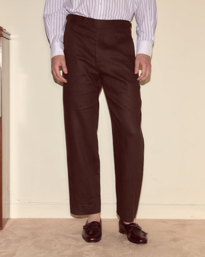 A person wearing Lightweight Linen Side-Buckle Trousers in Brown/Black, a white striped shirt, and black loafers stands against a cream wall.