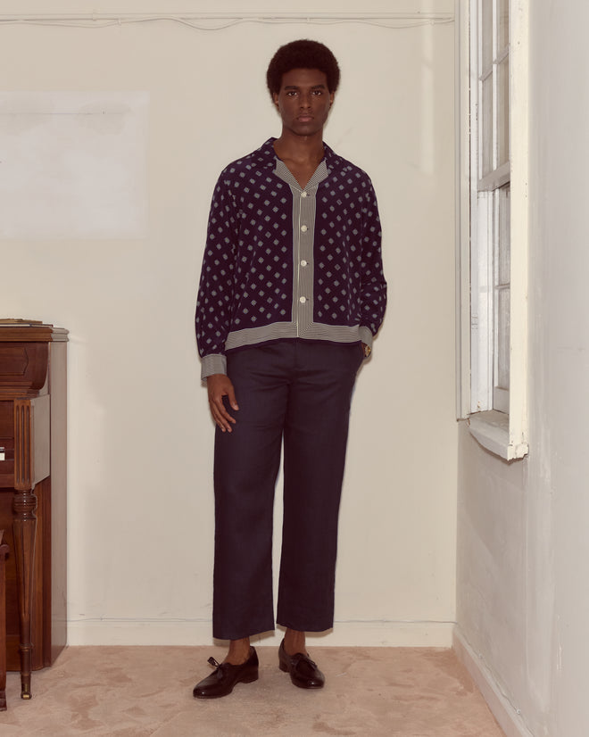 A man wearing Lightweight Linen Side-Buckle Trousers in Midnight stands indoors by a window, looking at the camera.