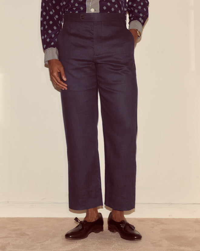 A person wearing Lightweight Linen Side-Buckle Trousers in Midnight, paired with a patterned shirt and black tassel loafers, stands against a plain wall.