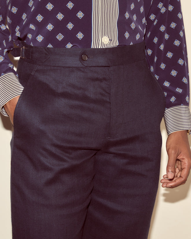 Person wearing Lightweight Linen Side-Buckle Trousers - Midnight, high-waisted with side-buckles, paired with a patterned shirt and one hand in their pocket.
