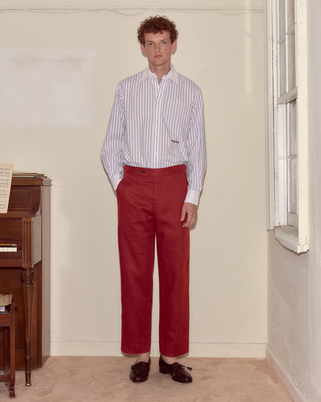 A man stands indoors beside a piano and window, wearing a striped shirt, loafers, and Lightweight Linen Side-Buckle Trousers in red.