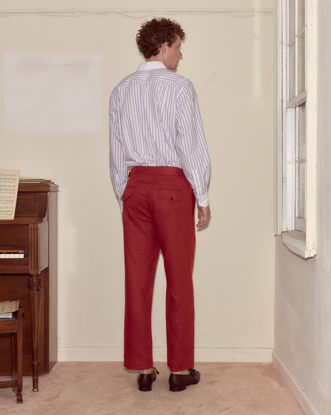 A man in a striped shirt and Lightweight Linen Side-Buckle Trousers - Red stands facing a beige wall near a piano and a window.