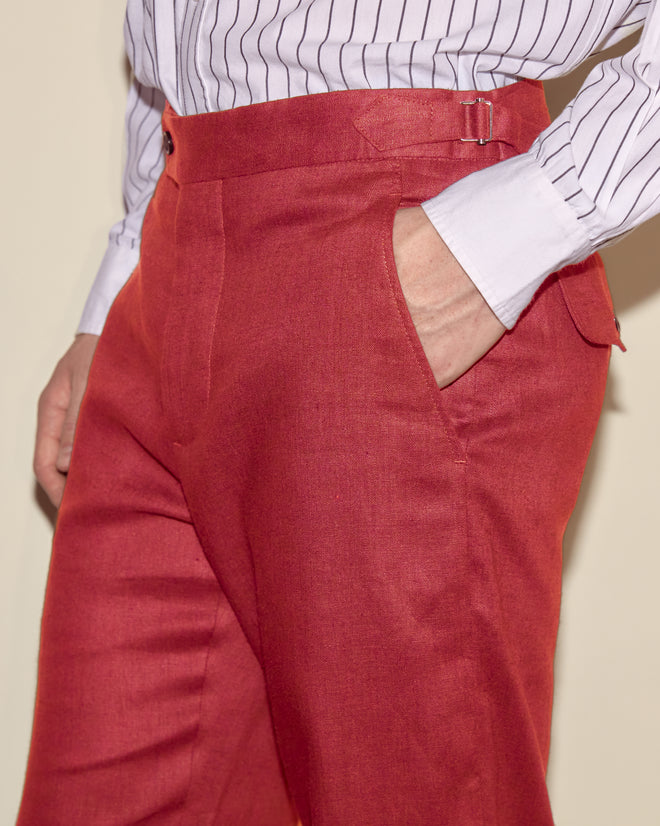 Model wearing Lightweight Linen Side-Buckle Trousers in red, paired with a white pinstripe shirt, hand in pocket; image shows view from waist to mid-thigh.