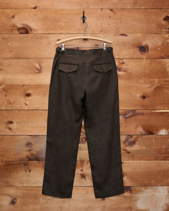 Lightweight Linen Side-Buckle Trousers in brown hang from a wooden hanger against a rustic wooden wall, viewed from the back—these men’s summer trousers offer a stylish and comfortable design.