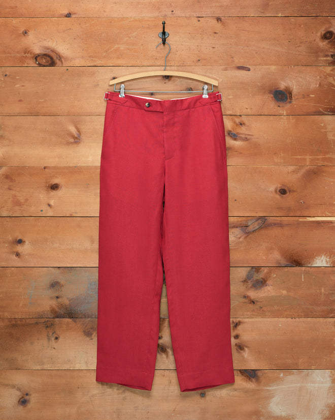 The Lightweight Linen Side-Buckle Trousers in red are displayed on a wooden hanger against a rustic wooden wall, highlighting an ideal choice for men’s spring trousers.