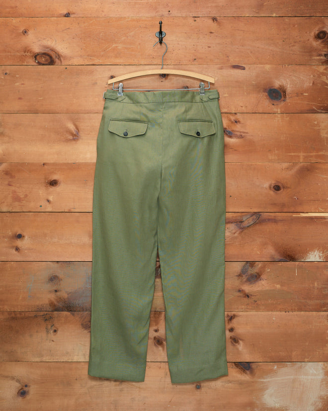 Lightweight Linen Side-Buckle Trousers in olive hang against a wooden wall background, providing a stylish and comfortable option for men’s summer wear.