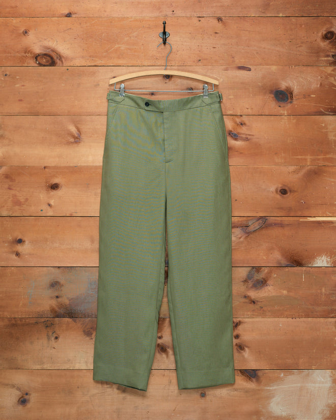 Lightweight Linen Side-Buckle Trousers - Olive are displayed on a wooden hanger against a rustic wooden wall. These men’s summer trousers combine classic style with practical side-buckle details, making them ideal for warm-weather wear.