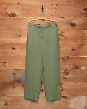 Lightweight Linen Side-Buckle Trousers - Olive are displayed on a wooden hanger against a rustic wooden wall. These men’s summer trousers combine classic style with practical side-buckle details, making them ideal for warm-weather wear.