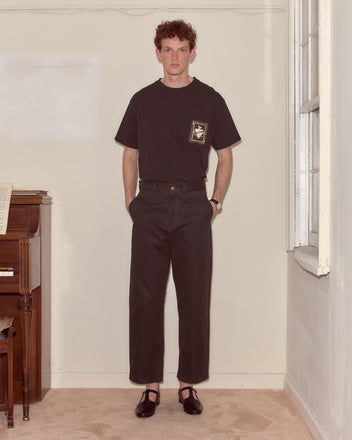 A man stands indoors in American collegiate style, wearing a black T-shirt, Campus Trousers - Black, and black shoes next to a piano and a window.