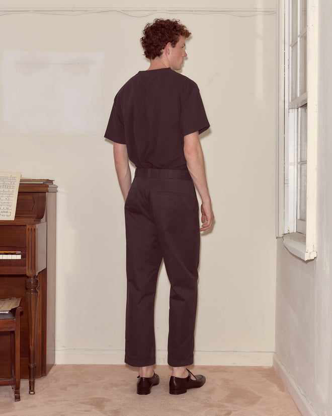 A man in Campus Trousers - Black made from heavyweight cotton twill stands facing a wall in a room with a piano and a window.