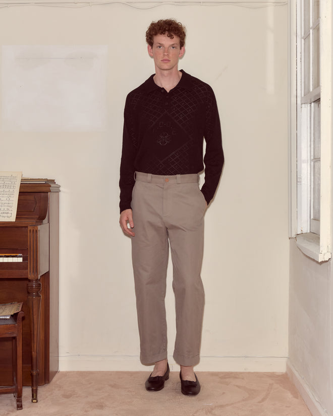 A man in a black sweater and grey Campus Trousers stands indoors by a piano, near a window on a carpeted floor, evoking classic collegiate style.