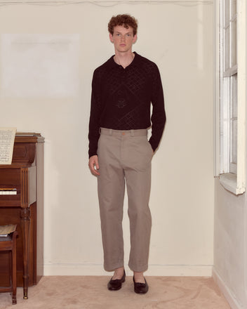 A man in a black sweater and grey Campus Trousers stands indoors by a piano, near a window on a carpeted floor, evoking classic collegiate style.