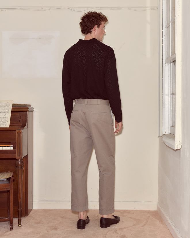A man stands indoors facing a wall, wearing a black shirt, Campus Trousers - Grey, and black shoes near a piano, channeling collegiate style.