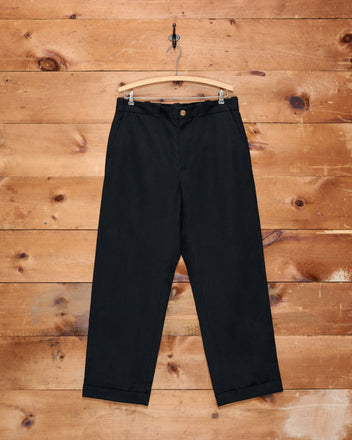 Campus Trousers - Black hang on a wooden hanger against a rustic wooden wall background. Crafted from heavyweight cotton twill, these high-waisted pants effortlessly embody an American collegiate style.