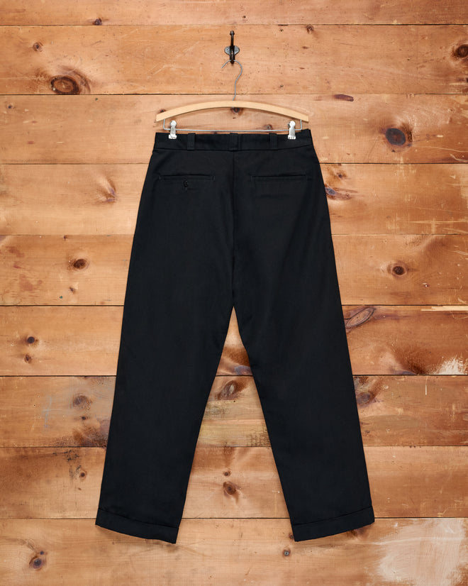 Campus Trousers - Black, crafted from heavyweight cotton twill, hang on a wooden hanger against a wooden wall display, showcasing their back pockets and cuffed hems—a nod to classic American collegiate style.