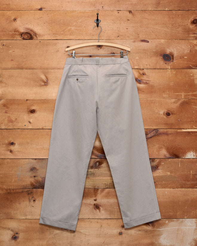 Campus Trousers - Grey, crafted from heavyweight cotton twill, hang on a wooden hanger against a rustic wooden plank wall, evoking classic American collegiate style.