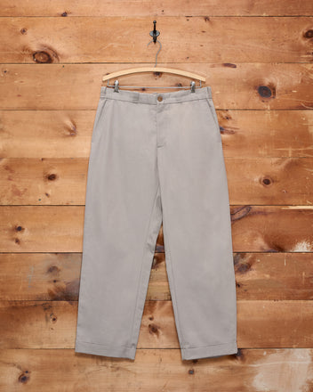 Campus Trousers - Grey, crafted from heavyweight cotton twill, hanging on a wooden hanger against a rustic wooden wall background, evoking a classic American collegiate style.