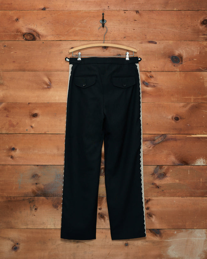 Lacework Side-Buckle Trousers crafted from black wool with scalloped machine lace trim, displayed against a wooden wall.