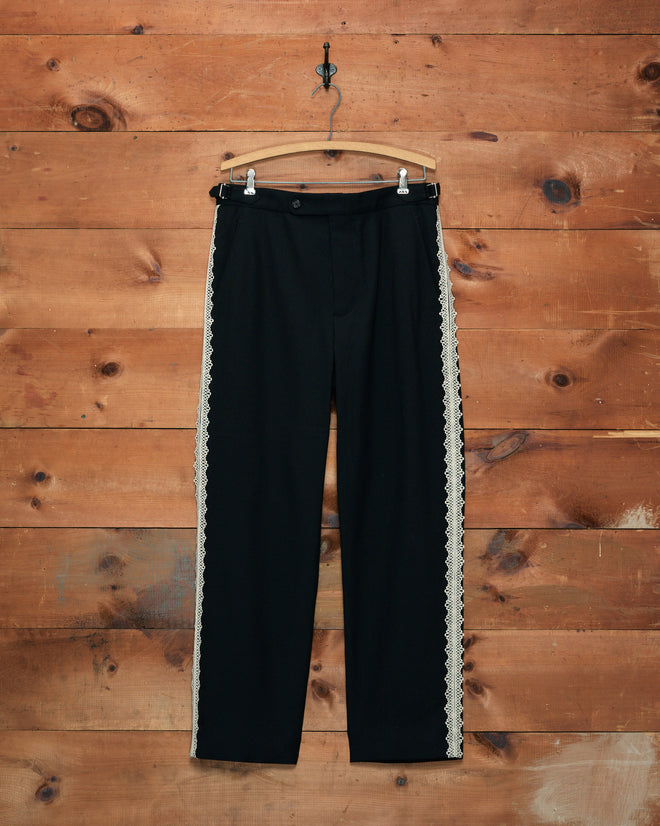 The Lacework Side-Buckle Trousers, crafted from black wool and featuring delicate white machine lace trim along the sides, are displayed hanging on a wooden wall using a hook.