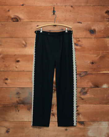 The Lacework Side-Buckle Trousers, crafted from black wool and featuring delicate white machine lace trim along the sides, are displayed hanging on a wooden wall using a hook.