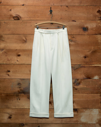 Off-white wool twill Riding McNab Trousers with pleated fronts and cuffed hems hanging on a wooden hanger.