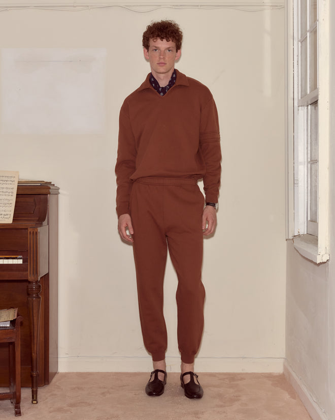 A man wearing the Crew Polo - Brown, crafted from 100% cotton in Portugal, stands indoors near a piano facing the camera with a neutral expression.