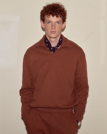 A man with curly red hair, dressed in a brown Crew Polo and a dark scarf, stands against a plain beige wall.
