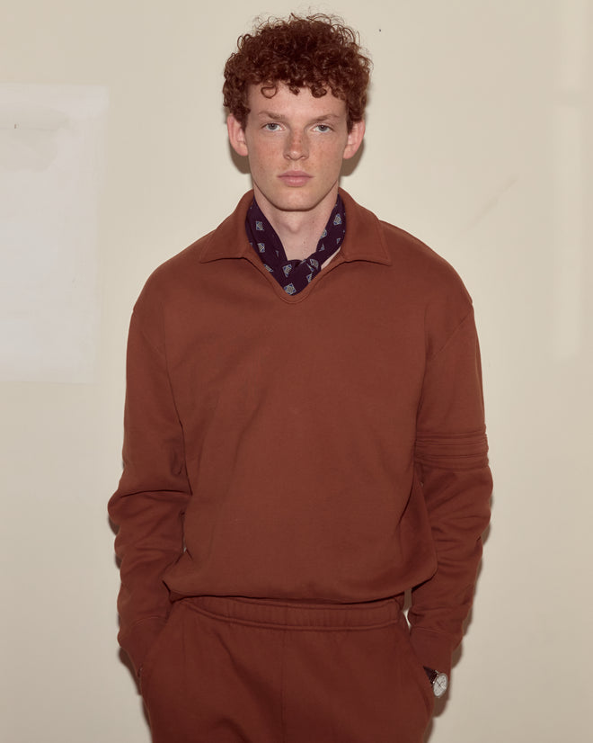 A man with curly red hair, dressed in a brown Crew Polo and a dark scarf, stands against a plain beige wall.