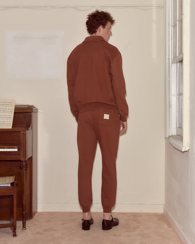 A man wearing the Crew Polo - Brown, made from 100% cotton, stands facing a wall next to a piano in a softly lit room.
