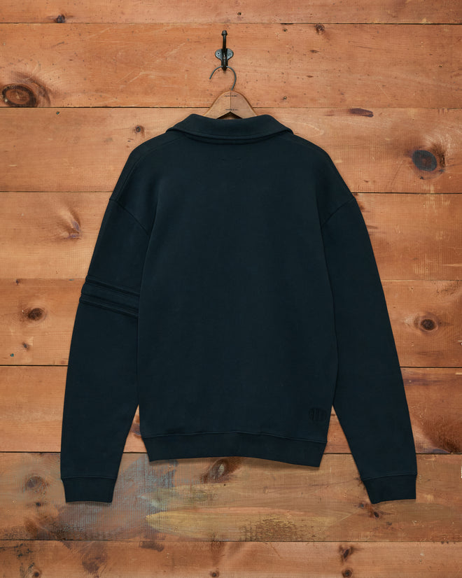 The black Crew Polo hangs on a hook against a wooden wall, viewed from the back. Made in Portugal from 100% cotton.
