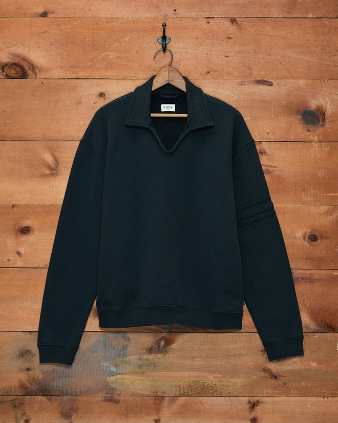 The Crew Polo - Black is a long-sleeve sweatshirt made in Portugal, shown on a hanger against a wooden wall background.