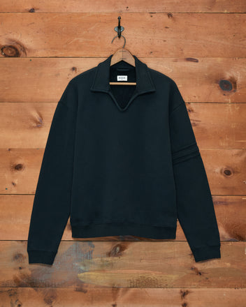 The Crew Polo - Black is a long-sleeve sweatshirt made in Portugal, shown on a hanger against a wooden wall background.