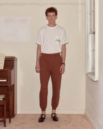 A man in a white t-shirt and brown joggers stands indoors near a piano and a window.