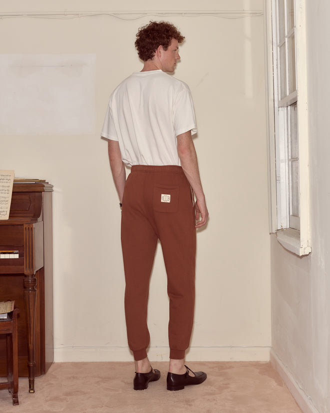 A man in a white shirt and brown joggers stands facing a beige wall near a window and a piano.