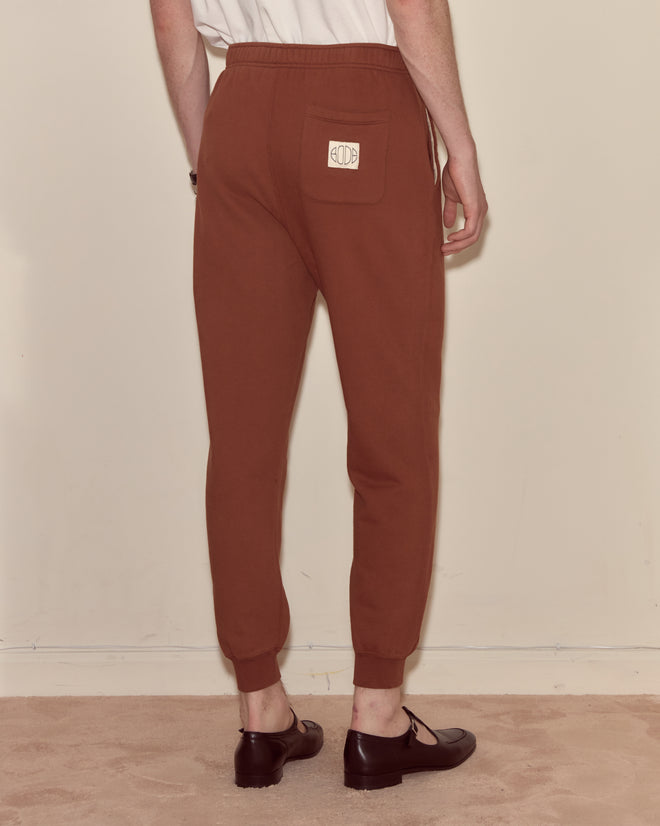 A person stands on a beige carpet facing away from the camera, wearing Joggers - Brown made from 100% cotton with a back pocket.