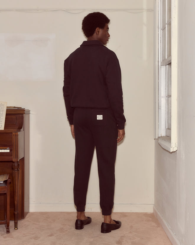 Man in black jacket and pants stands facing away in a beige room beside a piano and window, wearing the Crew Polo - Black, a classic cotton polo made in Portugal.