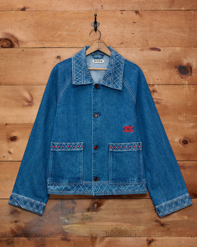 Embroidered Denim Jacket with white stitching and red detailing, hanging on a hook against a wooden wall—a classic piece of cotton outerwear that adds timeless style to any wardrobe.