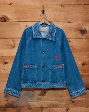 Embroidered Denim Jacket with white stitching and red detailing, hanging on a hook against a wooden wall—a classic piece of cotton outerwear that adds timeless style to any wardrobe.