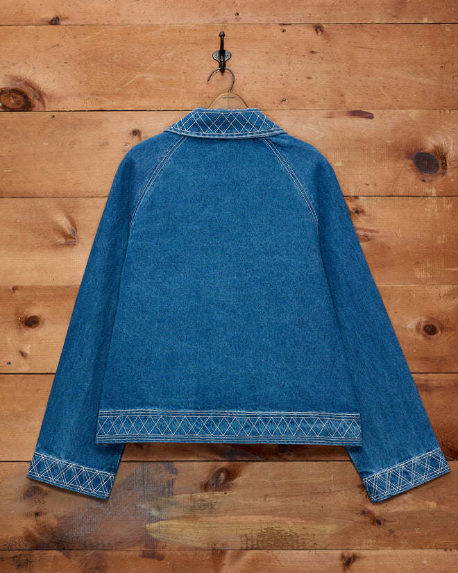 The Embroidered Denim Jacket, crafted from blue vintage denim with white stitching, is displayed on a hook against a wooden wall and shown from the back. This classic cotton outerwear showcases subtle details inspired by traditional embroidered workwear.