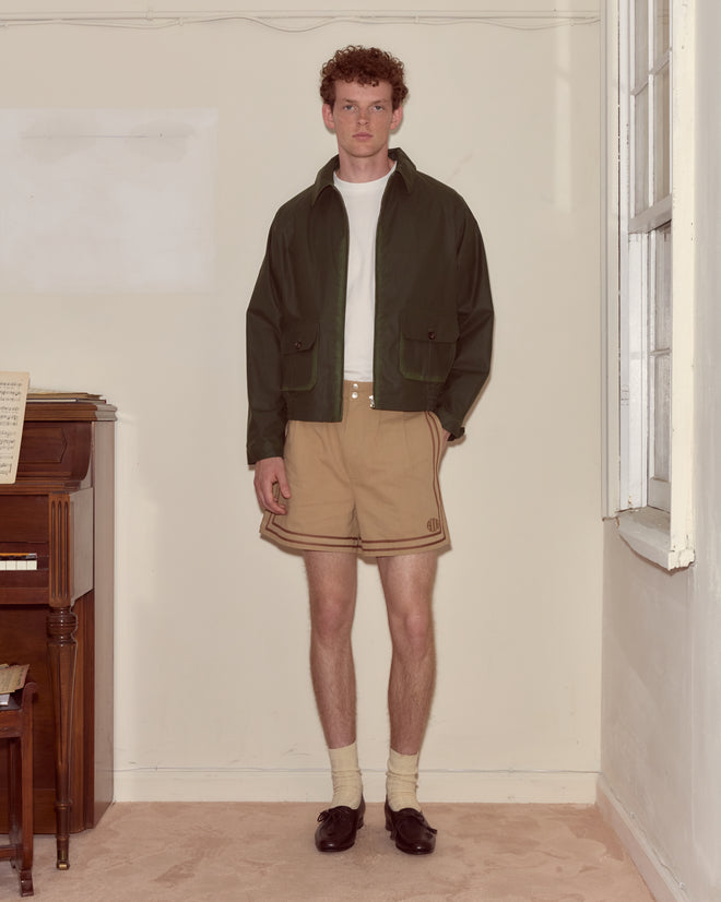 A man in a green Waxed Coach's Jacket stands indoors beside a piano and a window, wearing brown shorts and loafers.